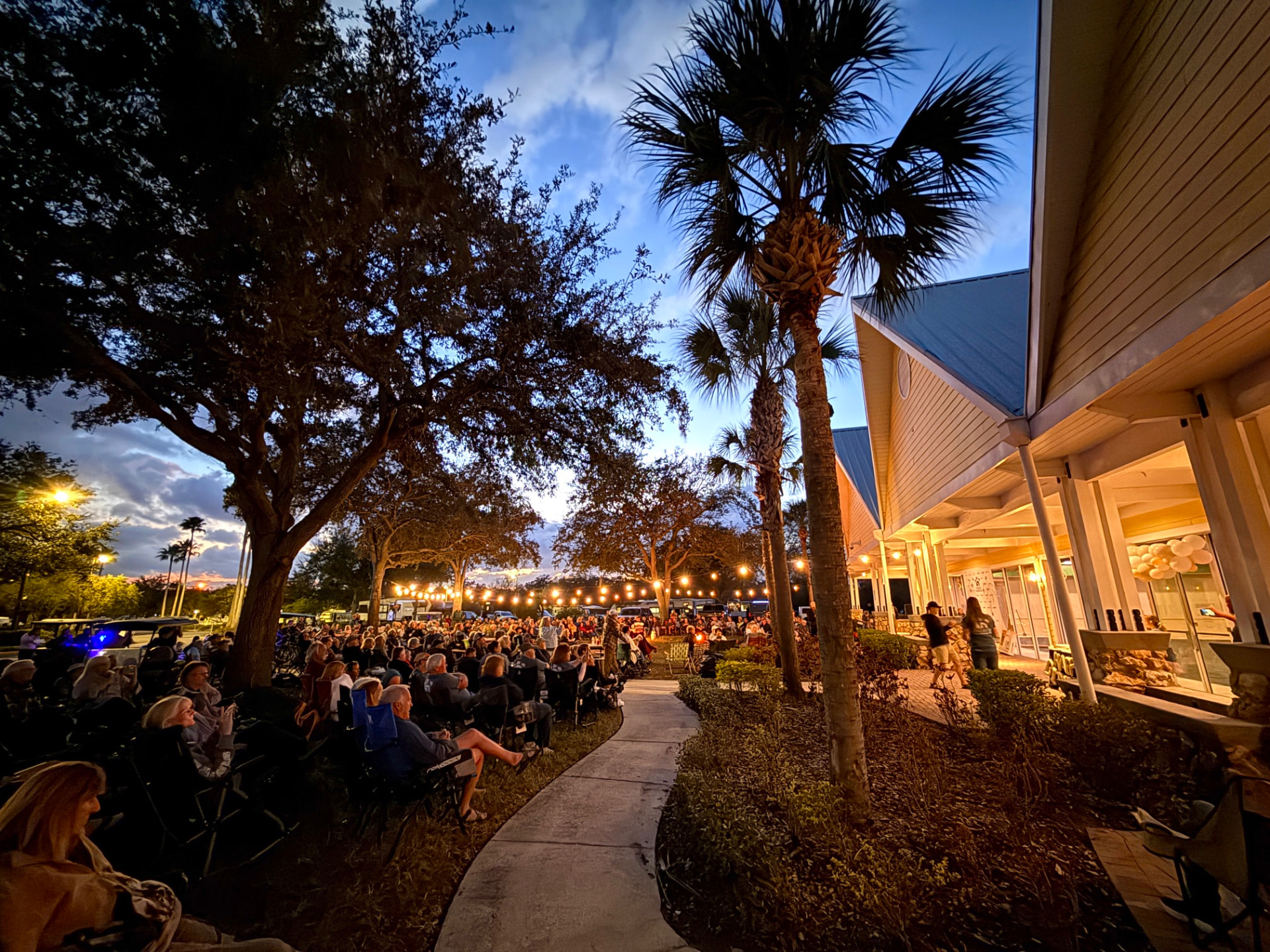 Crowd seated under palm trees and string lights for an evening concert